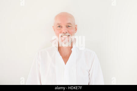 Alten Mann in Freizeitkleidung Blick in die Kamera Lächeln. Steht man vor der weißen Wand in seinem Haus. Fröhliche und positive Gesichtsausdruck zeigen. Stockfoto