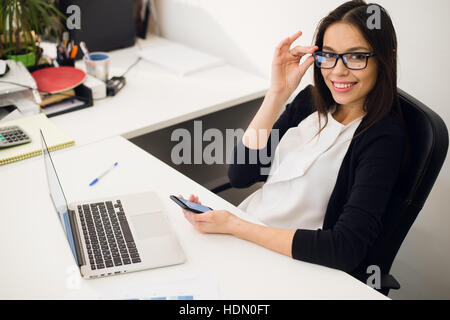 Ein gutes Geschäft zu sprechen. Fröhliche junge schöne Frau in Gläsern telefonieren mit Handy und Laptop mit Lächeln während Sie sitzen an ihrem Arbeitsplatz Stockfoto