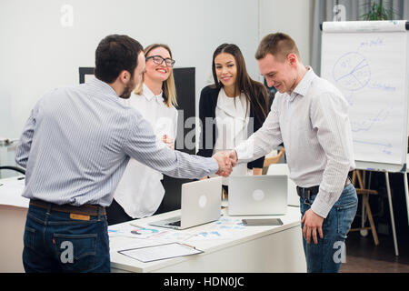 Willkommen Sie an Bord. Vier am Tisch während zwei von ihnen Geschäftsleute Händeschütteln und lächelnd Stockfoto