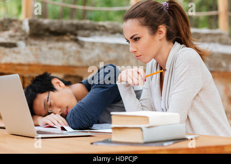 Konzentrierte sich die junge Frau sitzen und studieren in der Nähe von Menschen schlafen auf dem Schreibtisch Stockfoto