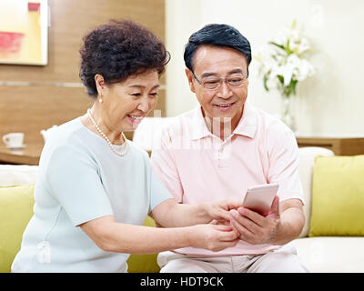 hochrangige asiatische paar sitzt auf couch Blick auf Handy zusammen, glücklich und lächelnd Stockfoto