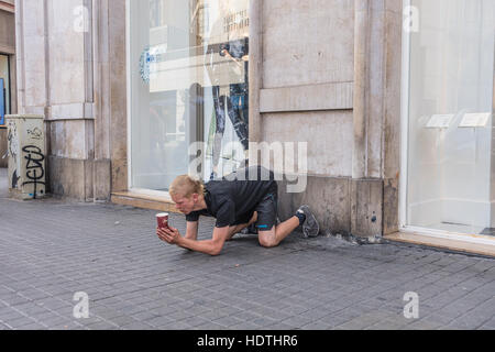 Ein 20er Jahre Männlich Bettler knelling auf einem Bürgersteig in der belebten Teil von Barcelona eine Tasse durchhalten, wie er bettelt. Stockfoto