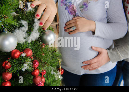 Schwangere paar in der Nähe von einem Weihnachtsbaum. Mann umarmt seine schwangere Frau. Mädchen schmückt einen Weihnachtsbaum. Stockfoto