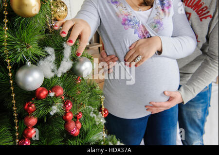 Schwangere paar in der Nähe von einem Weihnachtsbaum. Mann umarmt seine schwangere Frau. Mädchen schmückt einen Weihnachtsbaum. Stockfoto