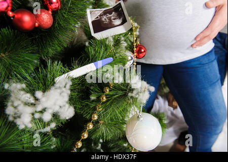 Ultraschallbild von Baby, Schwangerschaftstest am Weihnachtsbaum. Schwangere paar in der Nähe von einem Weihnachtsbaum. Mann umarmt seine schwangere Frau. Stockfoto