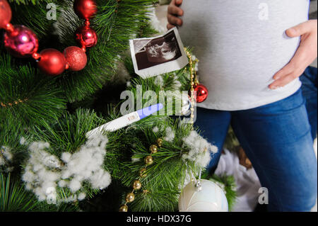 Ultraschallbild von Baby, Schwangerschaftstest am Weihnachtsbaum. Schwangere paar in der Nähe von einem Weihnachtsbaum. Mann umarmt seine schwangere Frau. Stockfoto