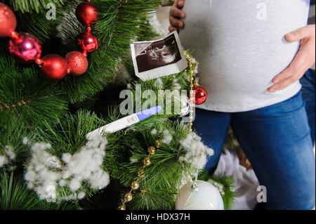 Ultraschallbild von Baby, Schwangerschaftstest am Weihnachtsbaum. Schwangere paar in der Nähe von einem Weihnachtsbaum. Mann umarmt seine schwangere Frau. Stockfoto