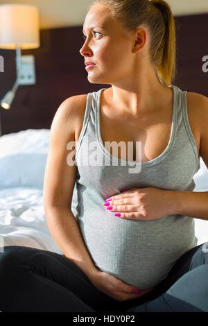 Nachdenkliche schwangere Frau im Bett mit der Hand am Bauch Stockfoto