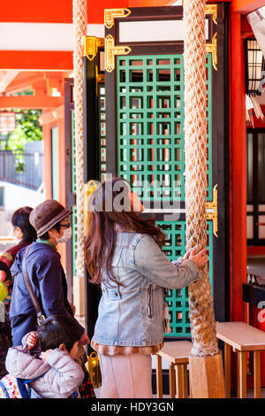 Japan, Kobe, Ikuta Shinto Schrein. Junge Frau beim Beten läutenden Glocke mit beiden Händen vor dem HONDEN. Stockfoto