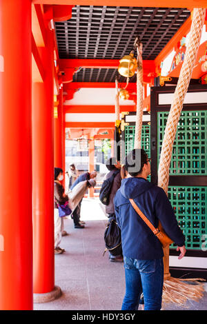 Japan, Kobe, Ikuta Shinto Schrein. Mann und Frau läutenden Glocke vor dem HONDEN. Vermillion Säulen und hängende Glocke Seile. Stockfoto