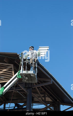 Ein männliche Dach Auftragnehmer in einem Boom Lift Korb tragen persönlicher Schutzausrüstung installiert Metall Dachplatten in einem Industriegebäude Stockfoto