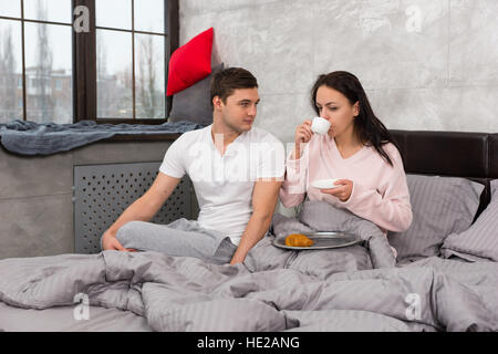 Junge Frau gerade aufgewacht, trinken eine Tasse Kaffee und ihr Frühstück im Bett mit ihrem Freund in der Nähe sitzen, tragenden Pyjamas Stockfoto