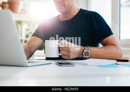 Bild des jungen Mannes mit Tasse Kaffee auf Laptop zugeschnitten. Geschäftsmann, arbeiten am Schreibtisch mit Kaffee. Auf Seite konzentrieren. Stockfoto