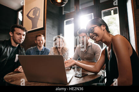 Lächelnde junge Frau, die etwas am Laptop an ihre Freunde. Gruppe von jungen Männern und Frauen im Café mit Blick auf Laptop-Computer und lächelnd.