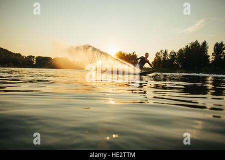 Wakeboarder bewegt sich schnell in Spritzwasser bei Sonnenuntergang. Man Wasserski an einem See. Stockfoto