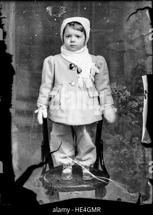 Porträt eines Kindes, das auf einem Stuhl sitzt, aufgenommen im Costică Acsinte Archiv, Ialomita, Rumänien. Dieses Foto aus einer alten Glasplatte repräsentiert das rumänische Leben des frühen 20. Jahrhunderts. Stockfoto