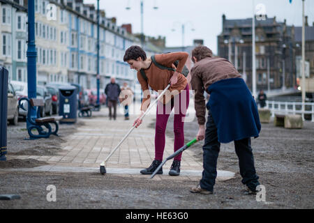 Aberystwyth Wales UK, Sonntag, 18. Dezember 2016 Aberystwyth University Studenten ELEANOR CLIFTON (22, Bürsten) und XANDER König (20, mit Schaufel), beide ursprünglich aus Wiltshire, habe alle von diesem Sonntagnachmittag Bürsten und Schaufeln Sand abseits die Promenade in Aberystwyth nach der stürmischen See Anfang dieser Woche. Sie haben sich auf eigene Faust, ohne Zutun der Gemeindebehörde Eleanors Partner zu ermöglichen, die ein Rollstuhl-Benutzer ist, und viele andere Leute eingeschränkten Mobilität, in den Genuss die Strandpromenade Credit: Keith Morris/Alamy Live-Nachrichten Stockfoto