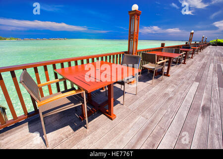 Stühle und Tische mit Palmen, Wasserbungalows und klarem Wasser auf luxuriöses Strandresort. Stockfoto