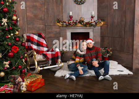 Süßes Paar in warme Pullover sitzt auf einem Teppich vor dem Kamin dekoriert und in der Nähe von Weihnachtsbaum mit präsentiert Stockfoto