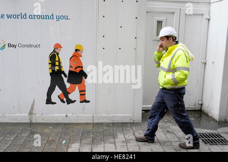 Ein Arbeiter chats auf seinem Handy, während er hinter einer temporären Wand zeigt Bilder der Arbeiter zu Fuß in die andere Richtung geht Stockfoto