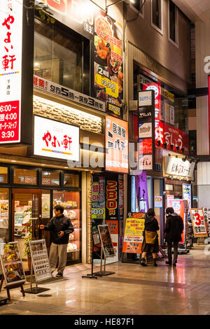 Japan, Kumamoto, Shimotori Arcade Street. Yayoi, bekannt als Yayoiken, Kette japanische Teishoku Restaurant bei Nacht mit anderen Geschäften. Einige Leute. Stockfoto