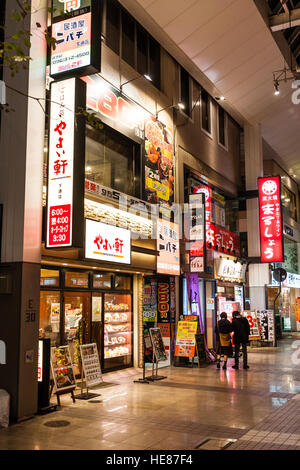 Japan, Kumamoto, Shimotori Arcade Street. Yayoi, bekannt als Yayoiken, Kette japanische Teishoku Restaurant bei Nacht mit anderen Geschäften. Einige Leute. Stockfoto