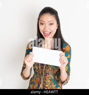 Porträt der jungen asiatischen Frau im traditionellen malaiischen Batik Kebaya Kleid Hand hält eine weiße leere Papierkarte mit überrascht Gesichtsausdruck, Stockfoto