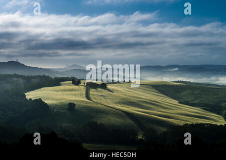 Typische grüne Toskana-Landschaft im Val d ' Orcia mit einem Bauernhof und eine kleine Kapelle auf einem Hügel, Felder, Zypressen, Bäume und am Morgen Nebel bei Sonnenaufgang Stockfoto