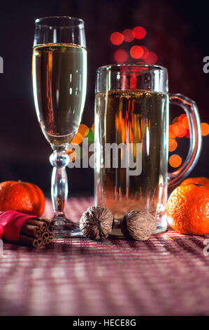 Silvester. Lustiges Bild ein Weinglas und ein Bierglas Champagner, Mandarine, Zimt und Walnüsse auf dem Hintergrund der Lichter Bokeh. Zurückhaltend. Stockfoto