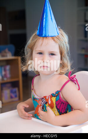 zwei Jahre alte blonde Kind mit leuchtend blauen Karton Partyhut und müde Facesitting in weißen Hochstuhl ernsthaft Stockfoto