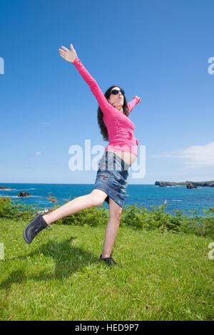 Brünette braune schwangere Frau mit rosa Trikot blau Jeansrock und schwarze Sonnenbrille glücklich tanzen auf grünen Rasen Wiese neben Ozean Stockfoto