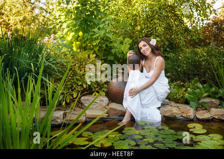 Schöne Mädchen in einem weißen Kleid hält einen Krug am Teich sitzen. Großen Tonkrug stand neben ihr. Stockfoto