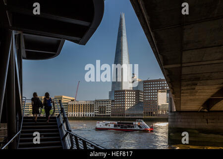 Zwei Frauen aufsteigend eine Treppe unter London Brücke mit einem Flussschiff und Shard Gebäude, London, Vereinigtes Königreich. Stockfoto