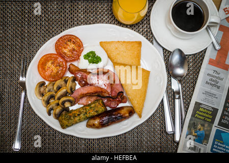 The Full Monty: A gebraten englisches Frühstück auf einem weißen Teller in einem Hotel UK (Speck, Ei, Wurst, Tomaten, Pilze, geröstetes Brot) Tasse Kaffee, Orangensaft und ein Newpaper Stockfoto