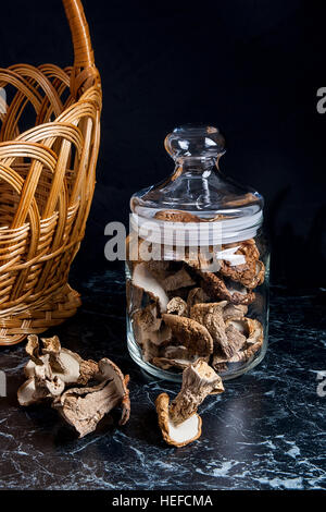 Trockene weiße Pilze im Glas und auf den Tisch. Gelb aus Holz Korb am hinteren Hintergrund. Komposition auf schwarzem Marmor Hintergrund Stockfoto