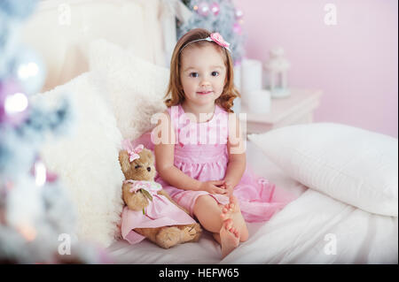 Sehr schöne charmante kleine Mädchen Blonde in rosa Kleid, sitzt auf einem Kinderbett und lacht laut den Hintergrund von Weihnachtsbäumen im hellen Innenraum des Hauses Stockfoto
