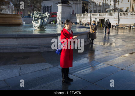 Eine asiatische Frau, gekleidet in einem leuchtend roten Mantel mit ihrem Handy am Londoner Trafalgar Square Stockfoto