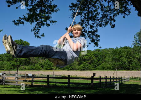 Ein 12-14 jähriger Junge Teenager schwingen auf Baum schwingen auf einem hellen, sommerlichen Tag, mit einem Zaun und Wald im Hintergrund. Stockfoto