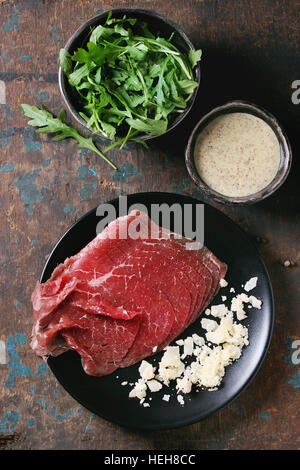 Zutaten für die Herstellung von Carpaccio. Geschnittenem Rindfleisch auf schwarze Platte, Senf und Parmesan-Sauce, Käse, Schüssel mit Rucola über alte dunkle Textur-Hintergrund Stockfoto