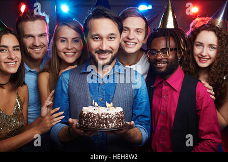 Attraktive asiatische Mann, hält Geburtstagskuchen mit brennenden Kerzen unter seinen interkulturellen Freunden Stockfoto