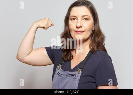 Mittlere gealterte und selbstbewusste Frau in Latzhosen ihrem Bizeps Muskeln, Zeichen für Selbstbewusstsein, hellgrauen Hintergrund Stockfoto