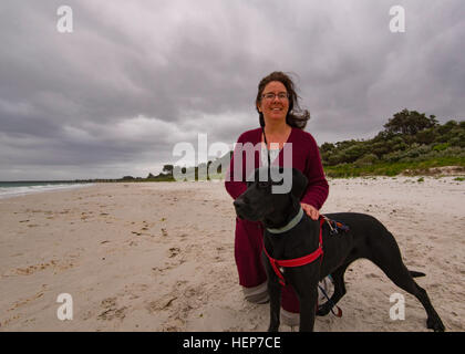 Eine Frau kniet neben einem schwarzen Hund in Callala Bay an einem windigen, bewölkten Tag in New South Wales, Australien Stockfoto