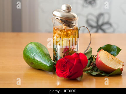 Teekanne mit Presse, eine rote Rose, eine Avocado, ein Stück Apfel auf dem Holztisch Stockfoto