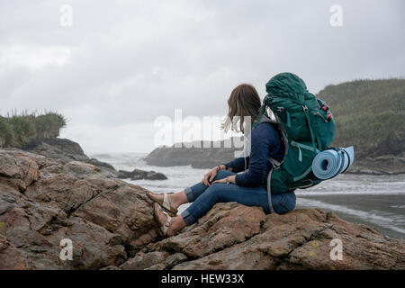 Junge Frau mit Rucksack sitzt auf Fels, konstante Bay, Charleston, Südinsel, Neuseeland Stockfoto