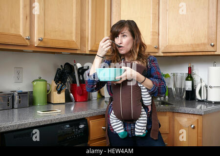 Mutter mit schlafenden Baby Boy in Baby-Tragetuch und Essen aus Schüssel Stockfoto