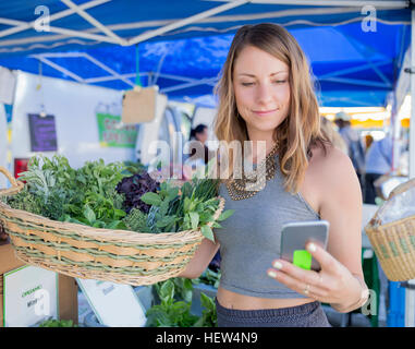 Frau an Obst und Gemüse Stand halten Korb mit frischen Kräutern Smartphone betrachten Stockfoto