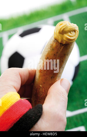 Hand, die Bratwurst Wurst mit Senf vor ein Fußballfeld mit einem Fußball, die deutschen Nationalfarben Stockfoto