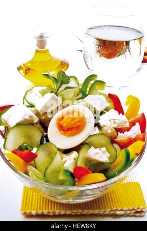 Griechischer Salat mit Feta-Käse, gekochtes Ei, Gurke, Tomaten und grünen und schwarzen Oliven und serviert mit einem Glas Weißwein Stockfoto