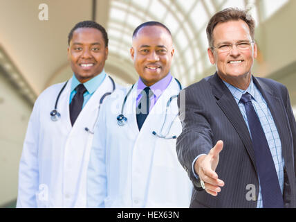 Zwei gemischte Rennärzte hinter Geschäftsmann für eine Hand Shake innen Krankenhaus zu erreichen. Stockfoto