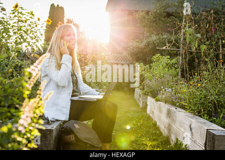 Frau mit Laptop sprechen auf Smartphone im sonnendurchfluteten Gemeinschaft Garten, Vancouver, Kanada Stockfoto